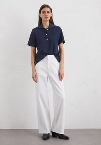 Marineblaues Poloshirt mit kurzen Ärmeln und Kragen sowie drei weißen Knöpfen, kombiniert mit weißen weit geschnittenen Hosen und schwarzen Absatzstiefeln.