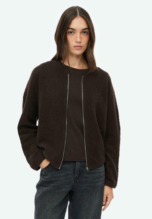 Frau mit langem braunem Haar, die einen dunkelbraunen Reißverschluss-Pullover über einem schwarzen Shirt und dunkel gewaschenen Jeans trägt, steht mit einer Hand in der Tasche.