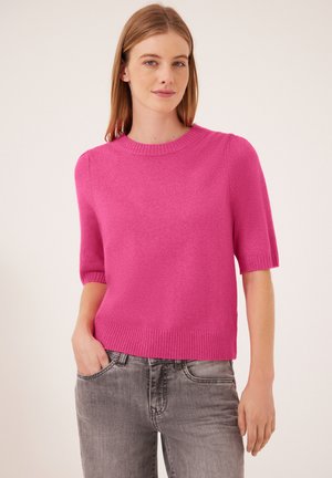Donna con capelli castano chiaro lisci che indossa un maglione rosa acceso a maniche corte e jeans grigi, che sta in piedi con una mano in tasca.