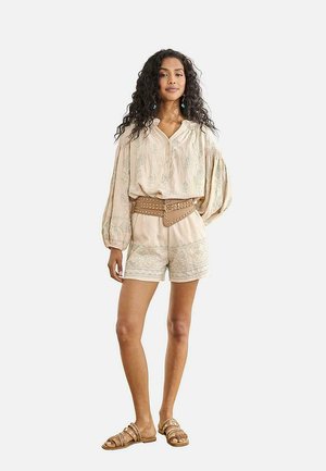Mujer con cabello largo y rizado, vistiendo una blusa beige bordada con mangas abullonadas, pantalones cortos a juego, cinturón ancho marrón con tachuelas y sandalias.