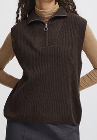 Pull sans manches côtelé marron avec un col montant et une fermeture éclair à moitié, associé à un haut beige ajusté à manches longues en dessous.