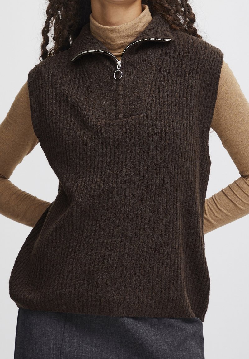 Pull sans manches côtelé marron avec un col montant et une fermeture éclair à moitié, associé à un haut beige ajusté à manches longues en dessous.