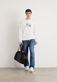 Biela mikina s logom "Calvin Klein", modré rifľové nohavice, čierna športová taška, biele tenisky, stojí na svetlosivých podlahe proti bielej stene.