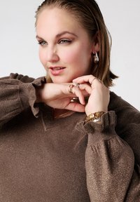 Bruine, gestructureerde trui met een V-hals en ruches aan de mouwen. Het model draagt meerdere ringen en een gouden armband met studs.