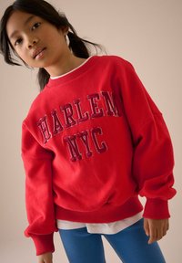 Rød sweatshirt med "HARLEM NYC" broderet i mørkerød, har oversized pasform og ribbede manchetter, parret med blå ribbede leggings.