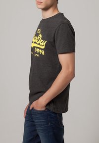 Camiseta de manga corta gris oscuro con texto gráfico amarillo en la parte frontal. Hecha de material suave, con un ajuste regular y escote redondo.