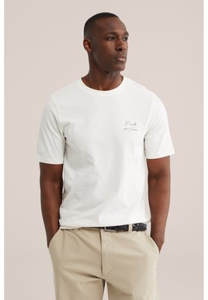 Muž s krátkymi vlasmi, ktorý má na sebe biele tričko s malým textom na hrudi a béžové nohavice, ruky v vreckách, pozerá sa doprava.