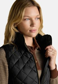 Blondynka w czarnej pikowanej kamizelce na chustę w brązowy wzór w ząb węża, trzymająca kołnierz kamizelki prawą ręką.