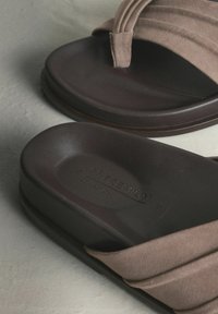 Sandales en daim marron avec une semelle lisse foncée, présentant un dessus plissé et un marquage minimal embossé sur la semelle intérieure.