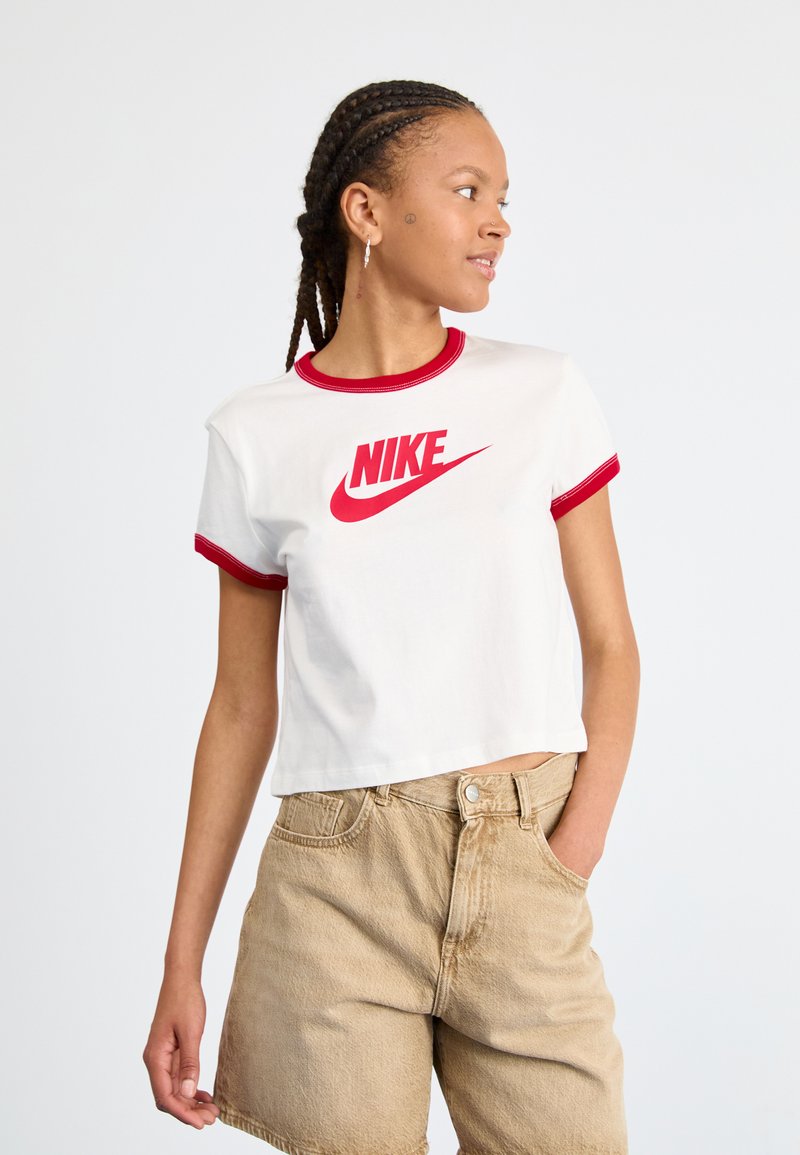 Valkoinen lyhythihainen t-paita, jossa on punaiset yksityiskohdat ja suuri punainen Nike-logo edessä, yhdistettynä beigekonvärisiin farkkushortseihin. Pehmeä puuvillakangas.