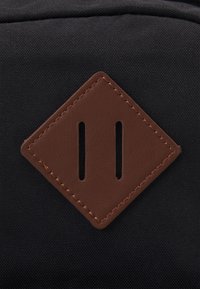 Patch en cuir marron en forme de losange avec deux fentes verticales cousues sur une surface en tissu noir.