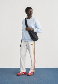 Νέος άνδρας με ανοιχτό μπλε φούτερ, λευκό παντελόνι φόρμας με πολύχρωμες ρίγες, κόκκινα αθλητικά παπούτσια, που κρατά μαύρη τσάντα χιαστί, στέκεται σε μπλε πάτωμα.