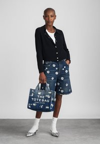 Torba tote z denimu z białym napisem "TORBA TOTE" i niebieskimi akcentami. Modelka ma na sobie czarny kardigan z prążkowanej tkaniny oraz dżinsowe szorty w kwiatowy wzór.