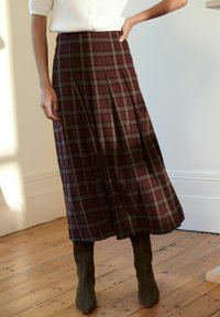 Jupe maxi écossaise bordeaux avec des plis et une coupe au genou, associée à des bottines en daim vertes. Présente une texture lisse et une forme structurée.
