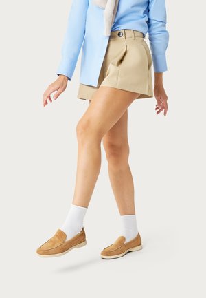 Persona vestida con pantalones cortos beige, zapatos tipo slip-on marrones con calcetines blancos y una camisa azul de manga larga sobre un fondo liso.