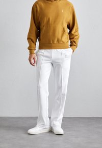 Felpa marrone con cappuccio e tasca frontale; abbinata a pantaloni bianchi a gamba dritta con cuciture frontali e dettaglio logo. Scarpe da ginnastica bianche.