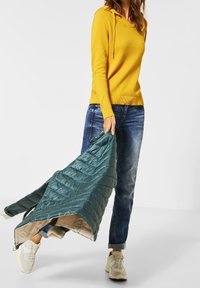Gelber Kapuzenpullover und blaue Jeans. Hält eine gesteppte türkisfarbene Jacke mit einem leichten Innenfutter, die ein puffiges, strukturiertes Design zeigt.