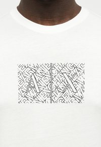 Λευκό πουκάμισο με μαύρο περιθώριο σε ορθογώνιο σχέδιο που εμφανίζει τα γράμματα "A" και "X" διαχωρισμένα με κάθετη γραμμή πάνω από διάσπαρτο μικρότερο κείμενο.