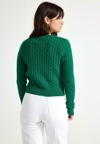 Woman facing sideways wearing a green cable-knit sweater and white pants against a plain light background.