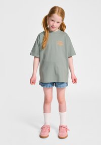 Grön bomullströja med rund halsringning och korta ärmar. I kombination med denimshorts. Skorna är rosa sneakers som kompletteras av vita strumpor.