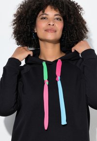 Donna con capelli ricci che sistema un felpa nera con lacci annodati multicolori verde, rosa e blu, indossando orecchini a cerchio.