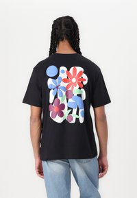 Sort t-shirt med et flerfarvet blomstermotiv på ryggen, med former i rød, blå og grøn mod en ensfarvet baggrund.