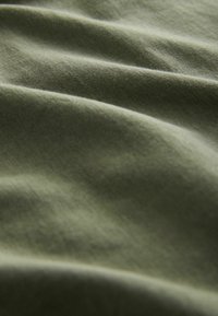 Olive zelena bombažna tkanina, ki prikazuje mehak teksturo z nežnimi valovi in gubami, ki poudarjajo njeno gladko površino in naravne barvne variacije.