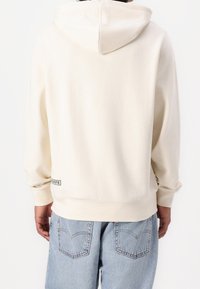 Krémová mikina s kapucí na stažení, žebrovanými manžetami a dolním okrajem, měkkou texturou a logem na spodní straně. Nosit s světle modrými džínami.