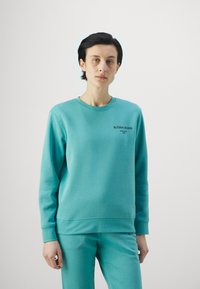 Sudadera en color teal de tejido suave, con mangas largas y cuello redondo. Logo "BJÖRN BORG" en negro en el pecho, combinada con pantalones de chándal a juego.