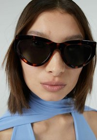 Cat-eye-solglasögon i mörkt sköldpaddsmönster med mörka linser. Tjocka bågar och spetsiga kanter, detaljrika med en subtil varumärkeslogotyp p å sidan.