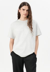 T-shirt en coton gris avec un col rond, des manches courtes et une coupe décontractée ; porté avec un pantalon noir. La texture semble lisse et douce.