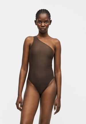 Vrouw die naar voren kijkt en een donkerbruine badpak met één schouder draagt, tegen een effen lichte achtergrond.