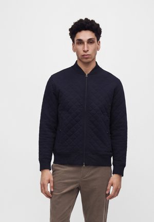 Homme portant une veste matelassée noire zippée et un pantalon marron, debout devant un fond clair uni.