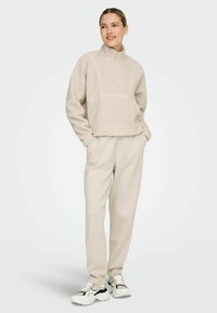 Beige fleece pullover with a high collar and quarter zip, paired with light beige sweatpants and chunky black-and-white sneakers.