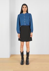 Chemise en denim à boutons avec des manches longues et bouffantes, associée à une jupe noire longueur genoux et des bottes de cowboy noires. Textures lisses et design ajusté.