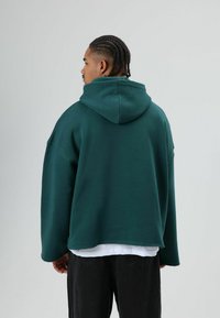 Übergroßer grüner Hoodie mit Kordelzugkapuze, strukturiertem Stoff und rohem Saum. Dazu schwarze Hose, Rückansicht.