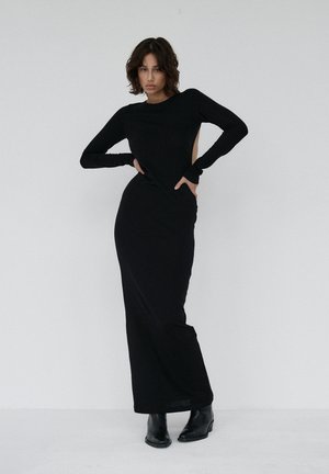 Kobieta pozująca w długiej czarnej dopasowanej sukience z długimi rękawami i otwartymi wycięciami po bokach, nosząca czarne botki, na jednolitym białym tle.