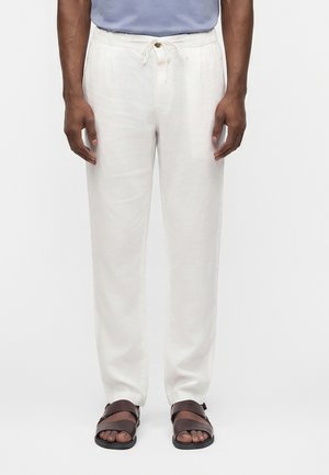 Uomo che indossa pantaloni bianchi larghi con coulisse e sandali marrone scuro, in piedi su uno sfondo chiaro e semplice.