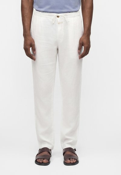 Homme portant un pantalon blanc ample avec un cordon et des sandales marron foncé, debout devant un fond clair uni.