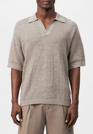 Hombre con camiseta tipo polo de punto de manga corta beige texturizada, con cuello abierto y pantalones beige a juego.