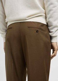 Koffiekleurige broek van een zachte stof, voorzien van twee achterzakken met knopen en een getailleerde pasvorm. De tailleband is bedekt door een crèmekleurige trui.