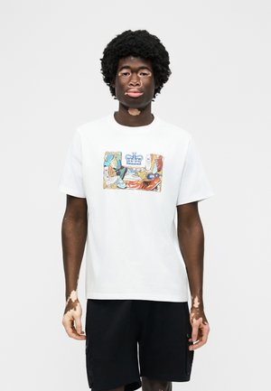 Άνδρας με λεύκη που φοράει λευκό μπλουζάκι με πολύχρωμο μοτίβο αθλητικού παπουτσιού και μαύρο σορτς, όρθιος μπροστά σε απλό φόντο.