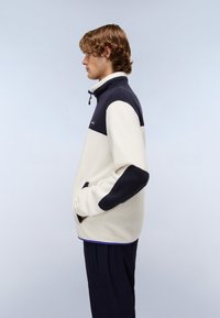 Giacca in pile color crema e blu navy, con colletto alto, chiusura a zip e toppe a contrasto sui gomiti. Tessuto liscio con vestibilità comoda.