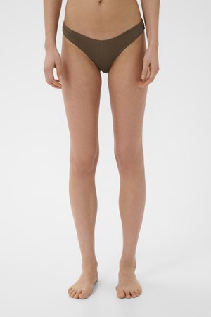 Frau steht barfuß, trägt olivgrüne Bikinihosen vor einfarbigem weißen Hintergrund, Frontansicht vom Taillenbereich bis zu den Füßen.