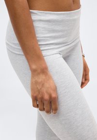 Κοντινό πλάνο ενός χεριού που ακουμπάει στον μηρό, φορώντας ανοιχτό γκρι εφαρμοστά παντελόνια και αντίστοιχη μέση, πάνω σε απλό φόντο.