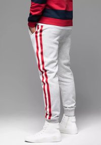 Graue Jogginghosen mit roten Seitenstreifen, Bündchen am Knöchel und seitlichen Taschen. Getragen mit weißen High-Top Sneakern. Lässiges, sportliches Design.