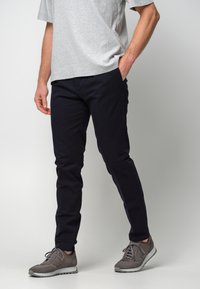 Dunkelblaue Hosen aus Baumwollmischgewebe, schlanker Schnitt mit Fronttaschen, kombiniert mit grauen Sportschuhen und einem grauen T-Shirt.