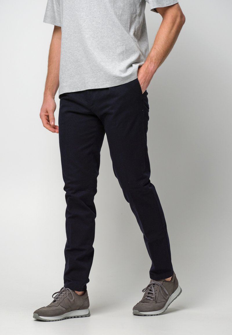 Dunkelblaue Hosen aus Baumwollmischgewebe, schlanker Schnitt mit Fronttaschen, kombiniert mit grauen Sportschuhen und einem grauen T-Shirt.