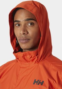 Chaqueta con capucha naranja y cuello alto, con un logo negro en el pecho y tela lisa, resistente al agua.