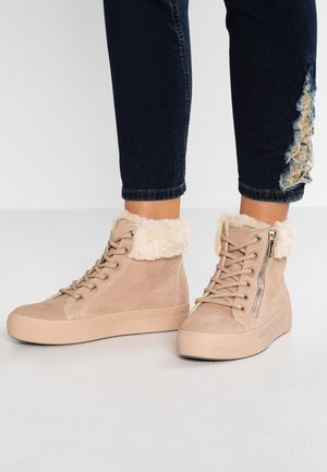 Baskets montantes beige avec bordure en fourrure et fermeture éclair latérale portées avec un jean skinny déchiré foncé sur une surface claire et plate.
